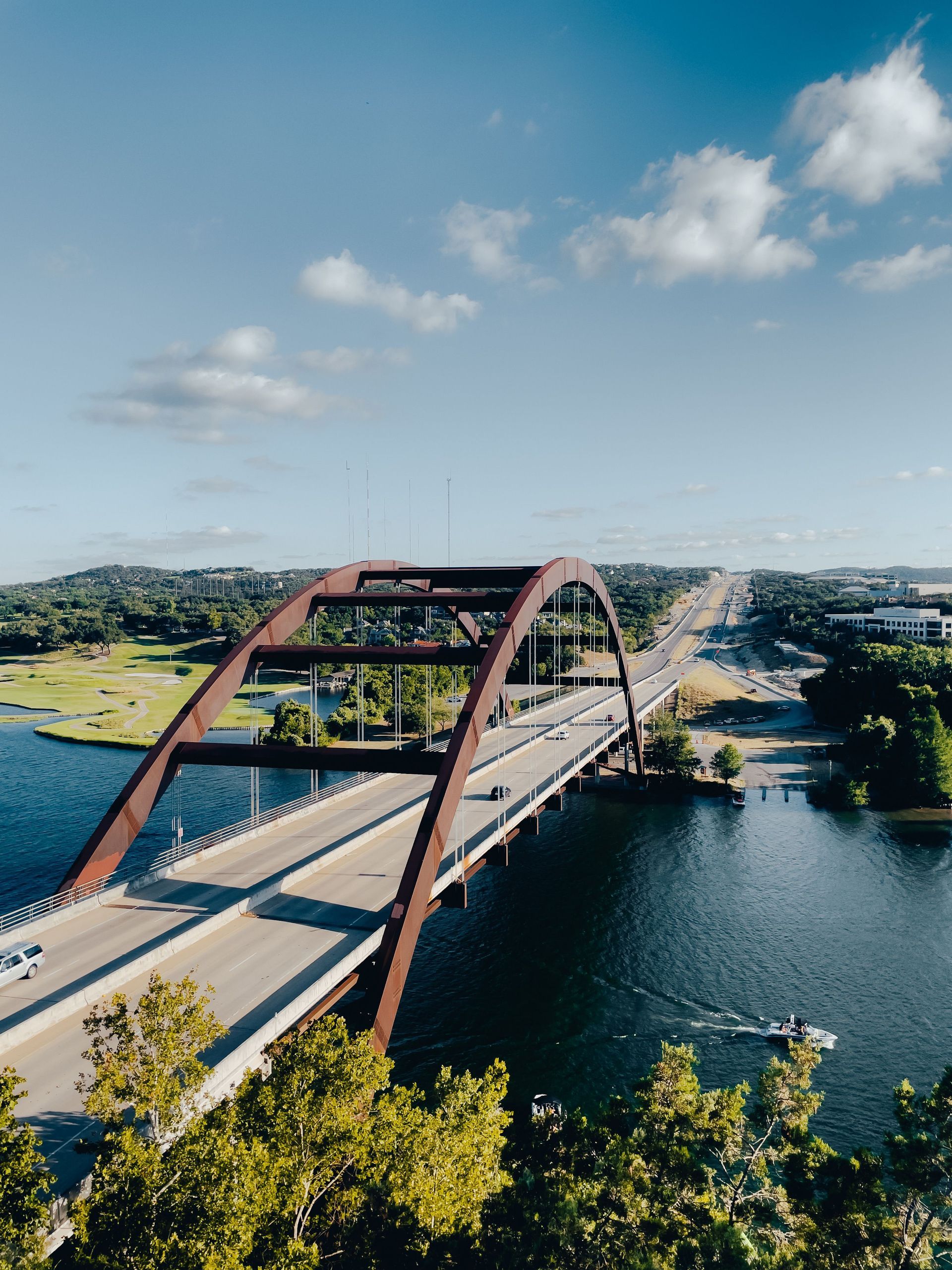 A red steel arch bridge spans a river under a blue sky, with green trees and shoreline nearby.