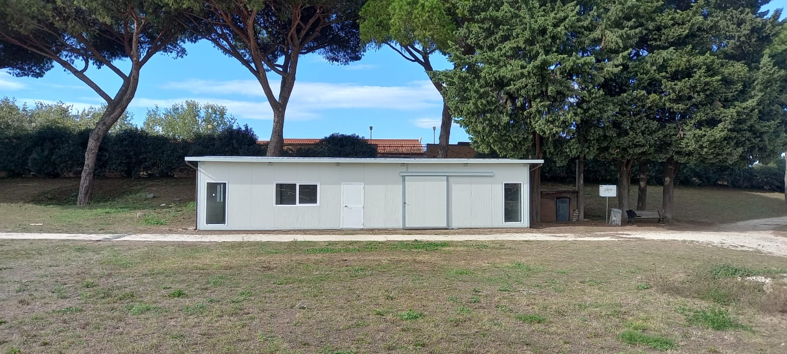 Edificio bianco con finestre e porta scorrevole, alberi e un campo erboso di fronte.