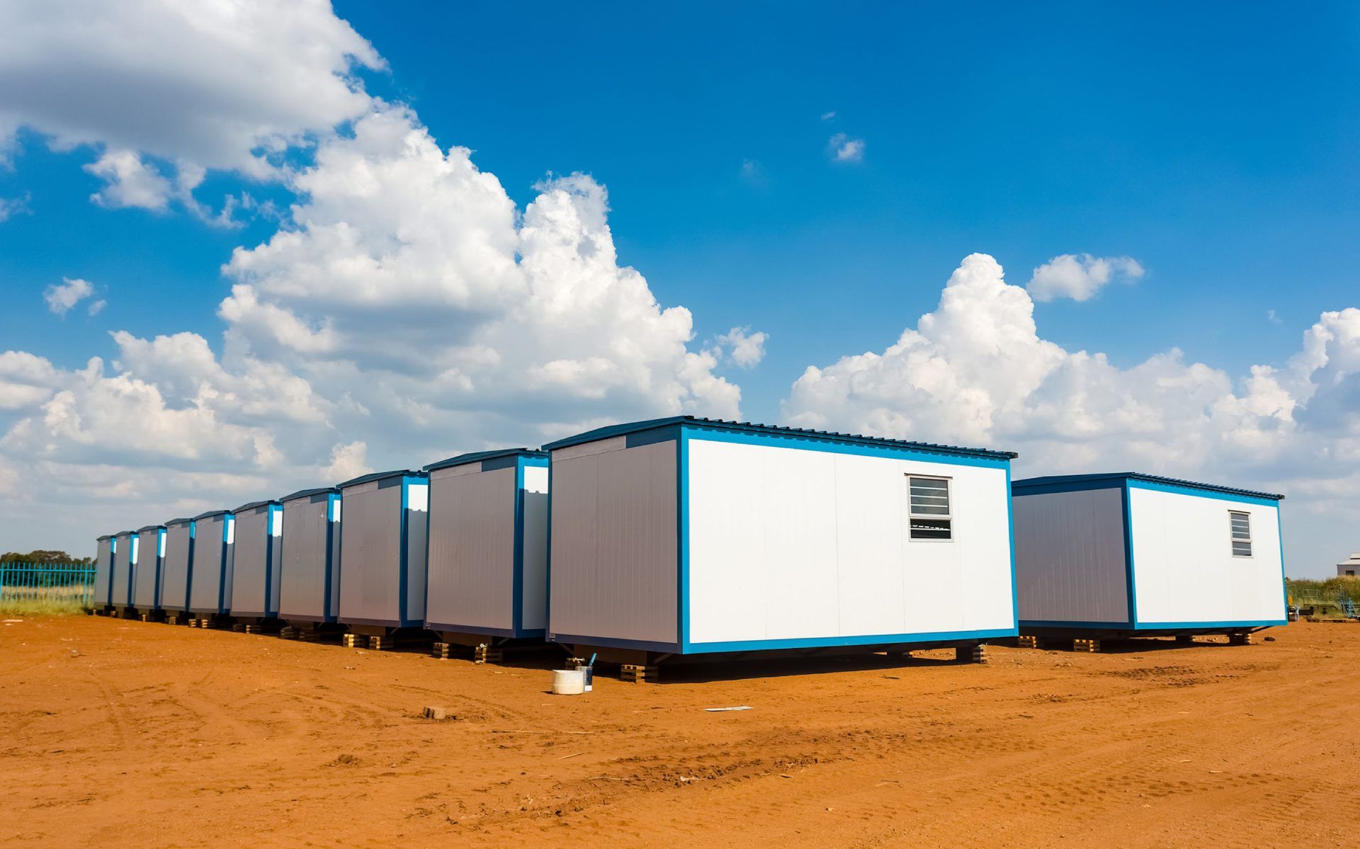 Fila di edifici portatili bianchi e blu su un campo di terra marrone sotto un cielo azzurro con nuvole.