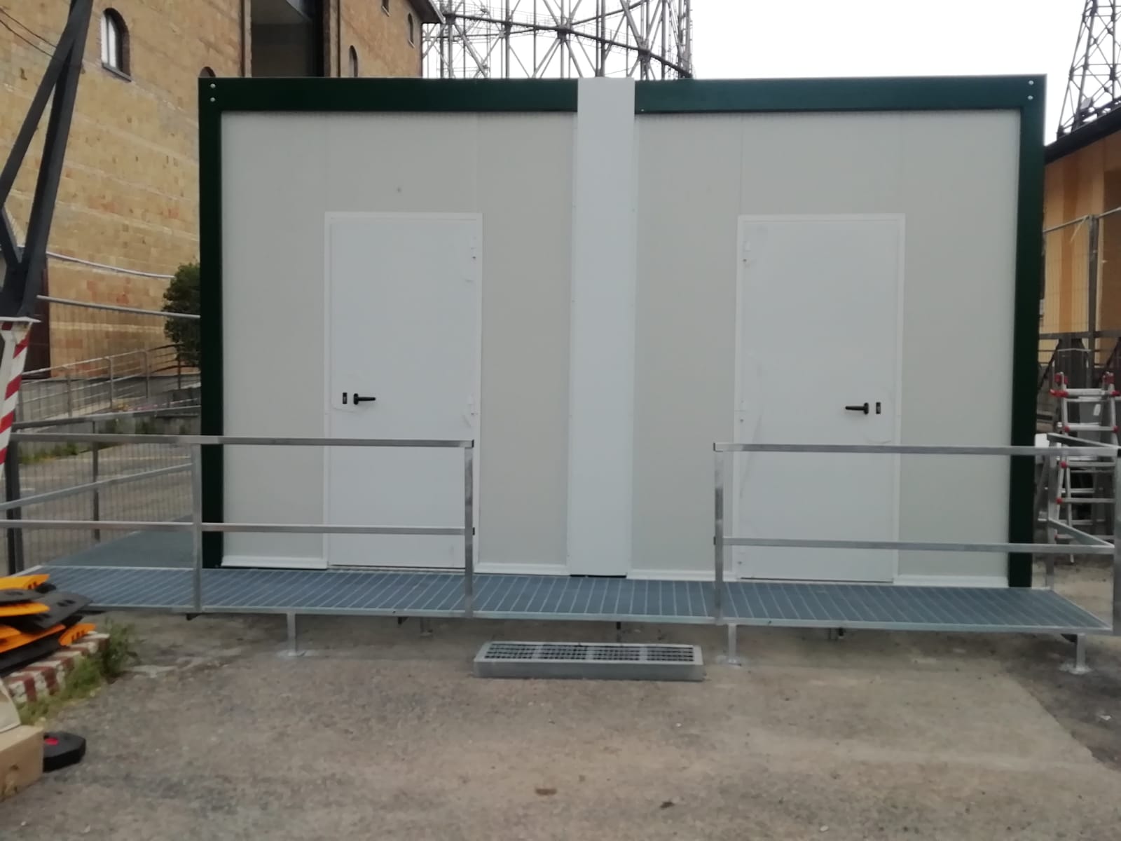 Due strutture mobili con pareti bianche e porte, collegate da una passerella metallica con ringhiere. Esterno, cantiere.