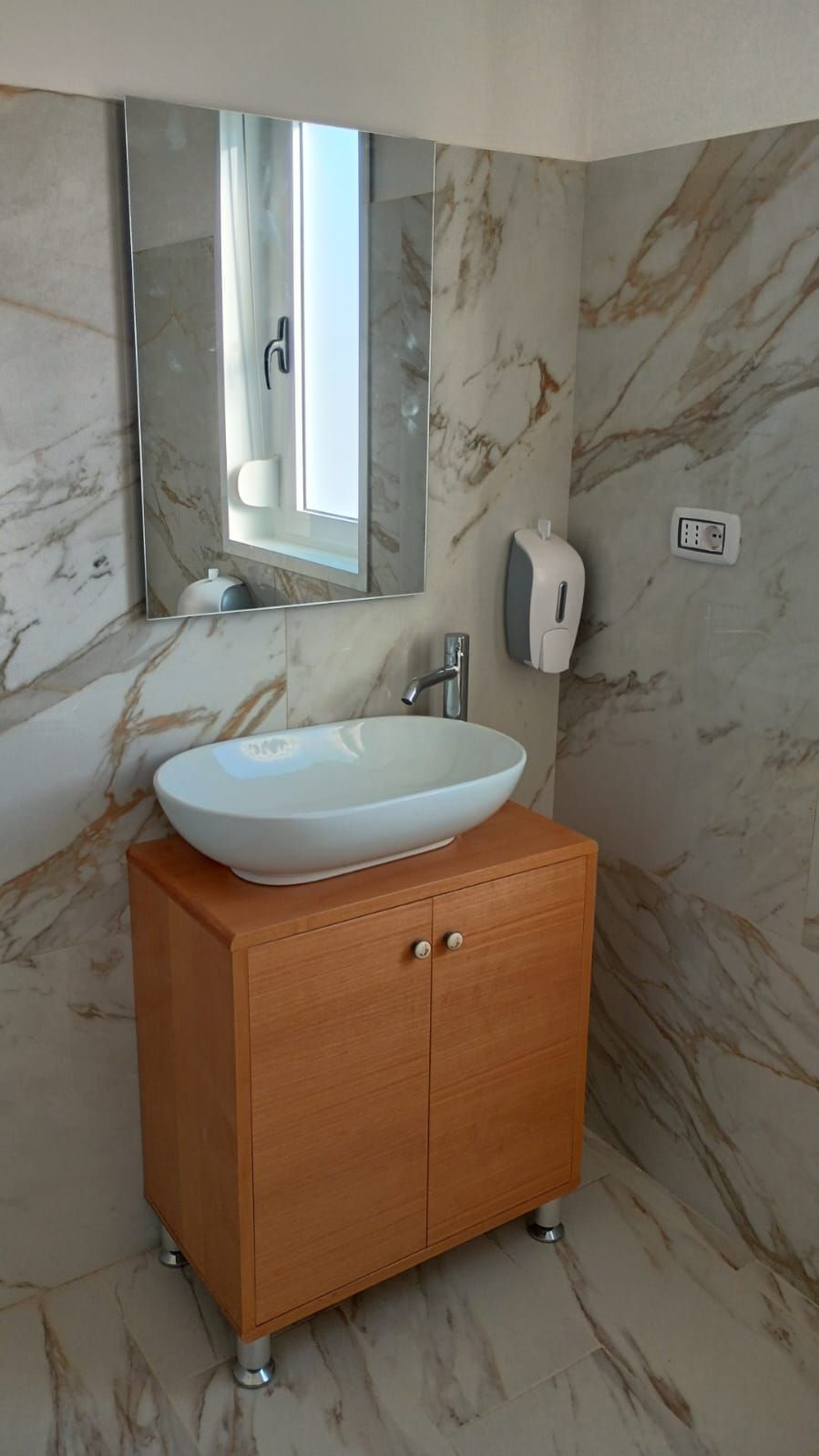 Bagno con mobiletto in legno, lavandino ovale bianco e un grande specchio su pareti in marmo.