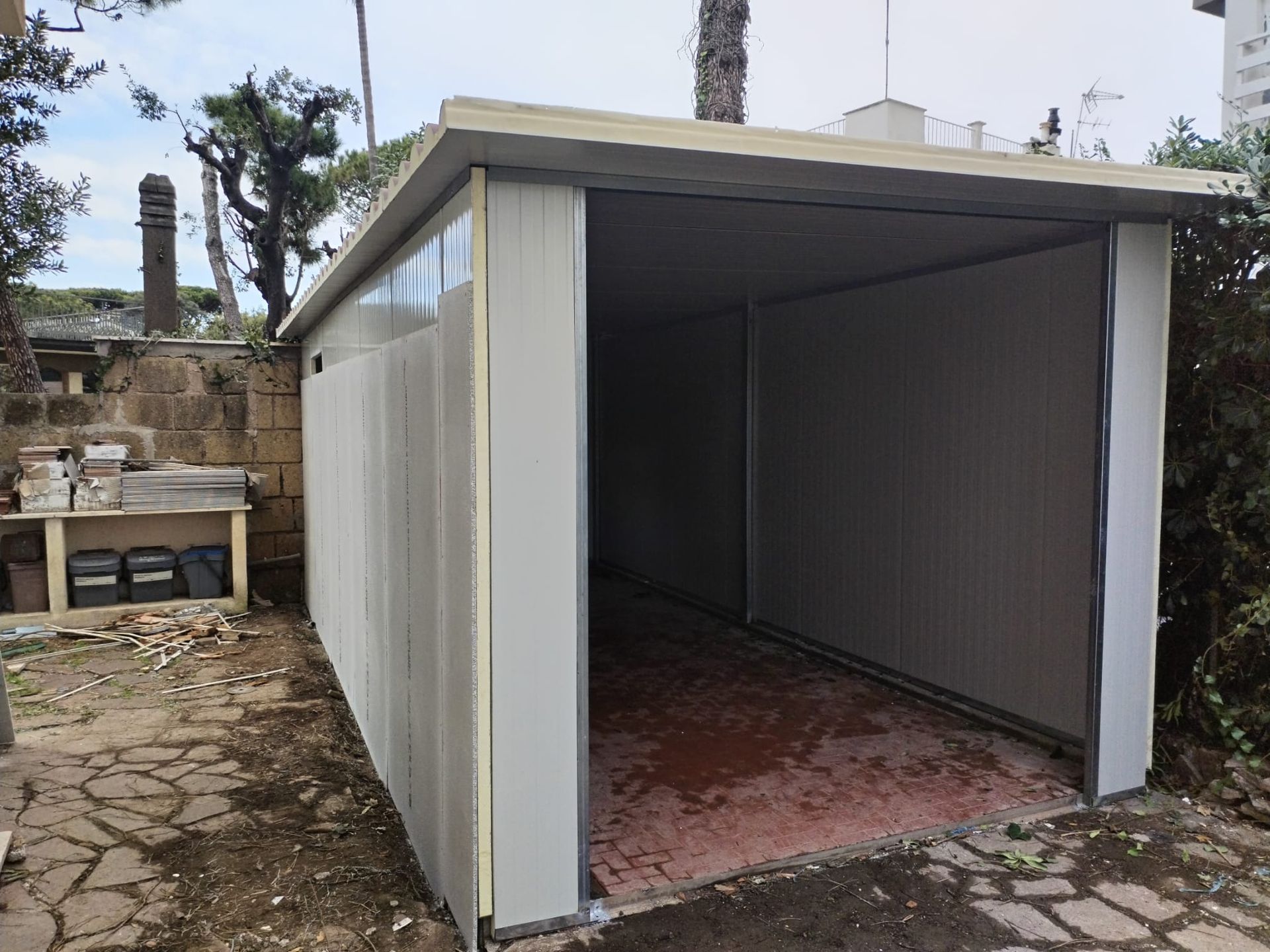 Un garage metallico con una porta aperta in un ambiente esterno. Le pareti sono grigie e il pavimento è rosso-marrone.
