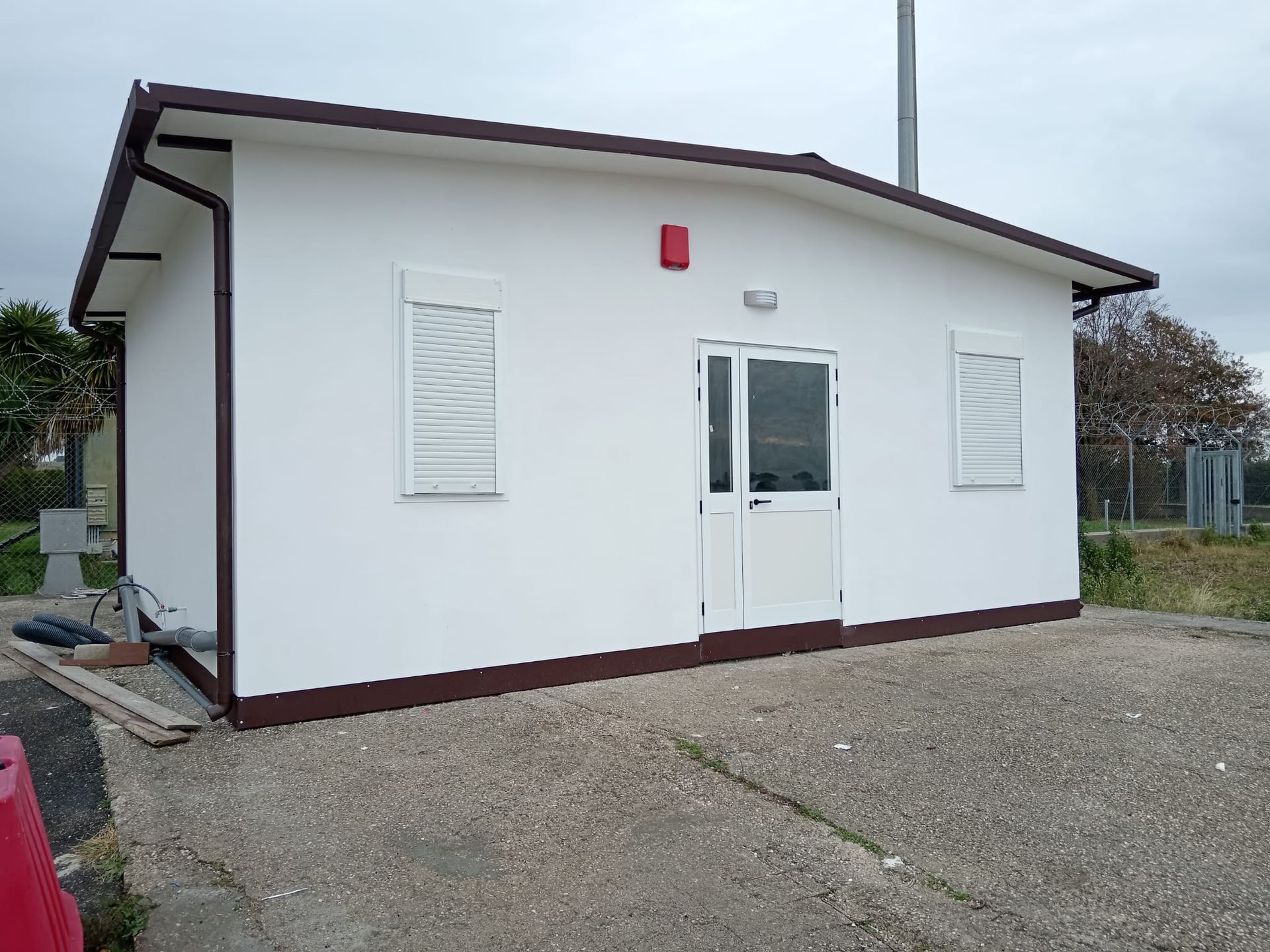 Edificio rettangolare bianco con finiture marroni, porta e due finestre con persiane su una superficie di ghiaia.