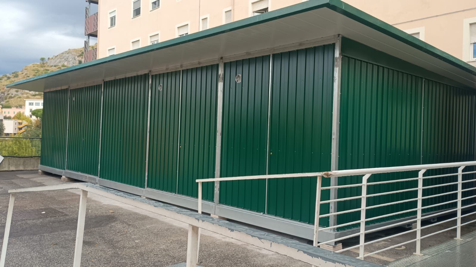 Capannone in metallo verde con tetto bianco su una piattaforma di cemento, accanto a una ringhiera.