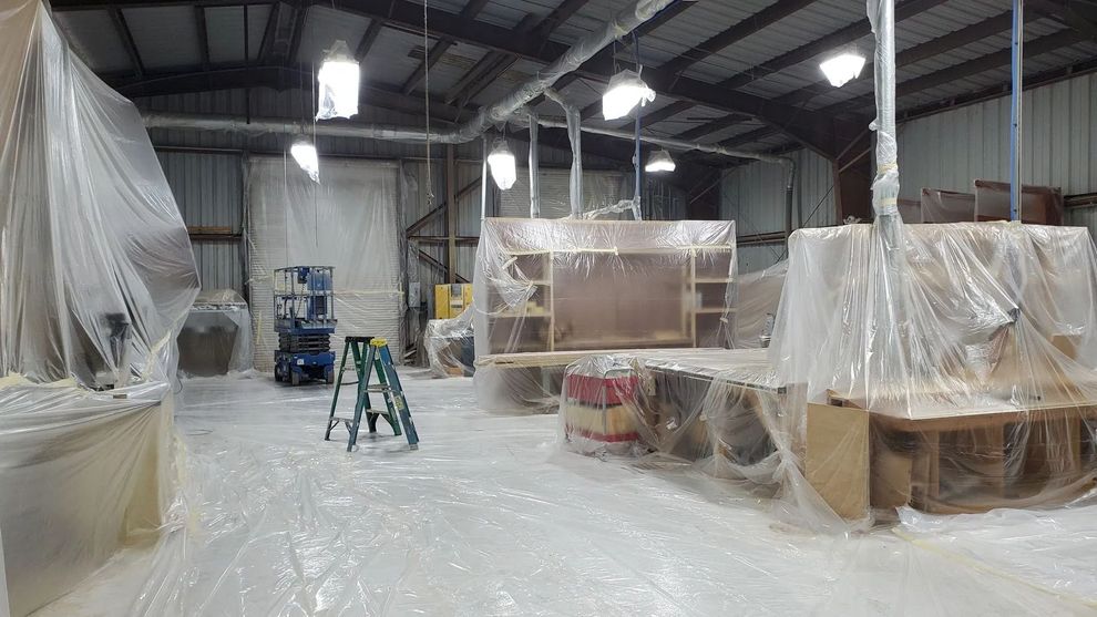 Warehouse interior with furniture covered in plastic for painting. Ladder and equipment visible.