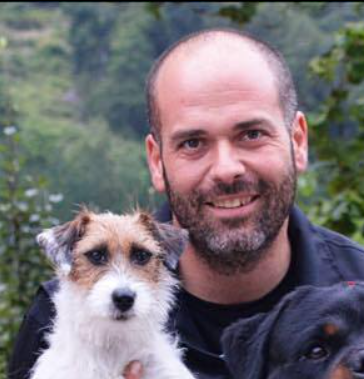 Un homme souriant tient un petit chien blanc et marron, ainsi qu'une partie d'un chien noir. En arrière-plan : des arbres.