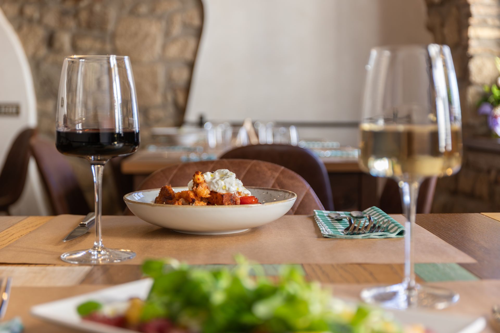 Calici da vino e un piatto di cibo su un tavolo di ristorante, con vegetazione sfocata in primo piano.
