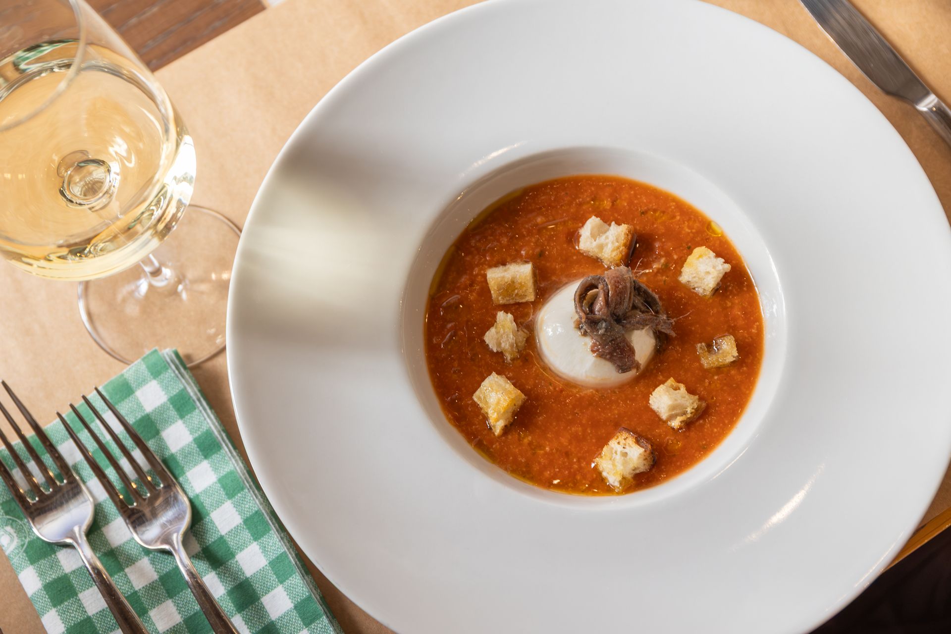Una ciotola bianca di zuppa rossa con crostini su un tavolo accanto a un bicchiere di vino bianco e un tovagliolo a quadretti verdi.