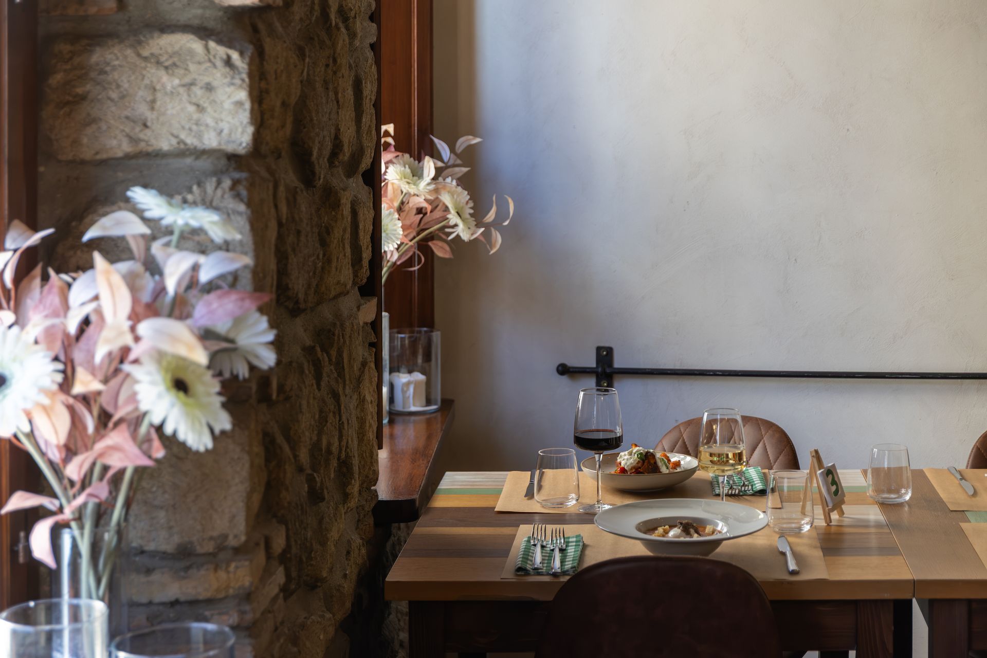 Tavolo da ristorante rustico apparecchiato con fiori, bicchieri e piatti accanto a un muro di pietra e a una zona salotto con finestre.