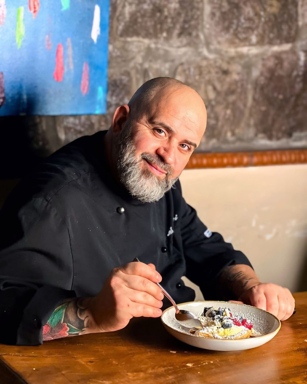 Un uomo con una camicia nera sorride mentre mangia un dolce a un tavolo di legno al chiuso.