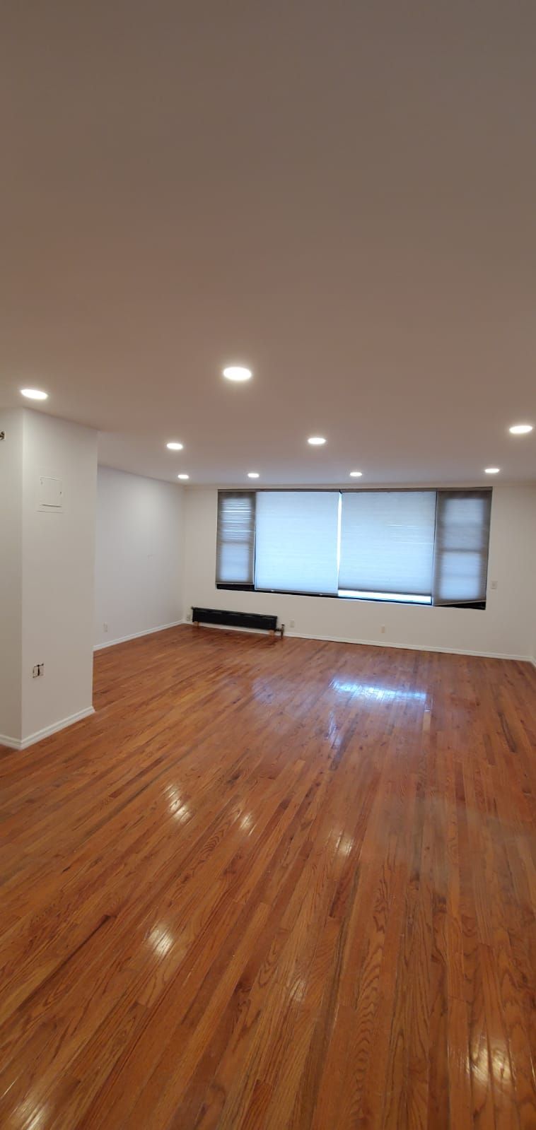 Empty living room with hardwood floors, recessed lights, and large windows with blinds.
