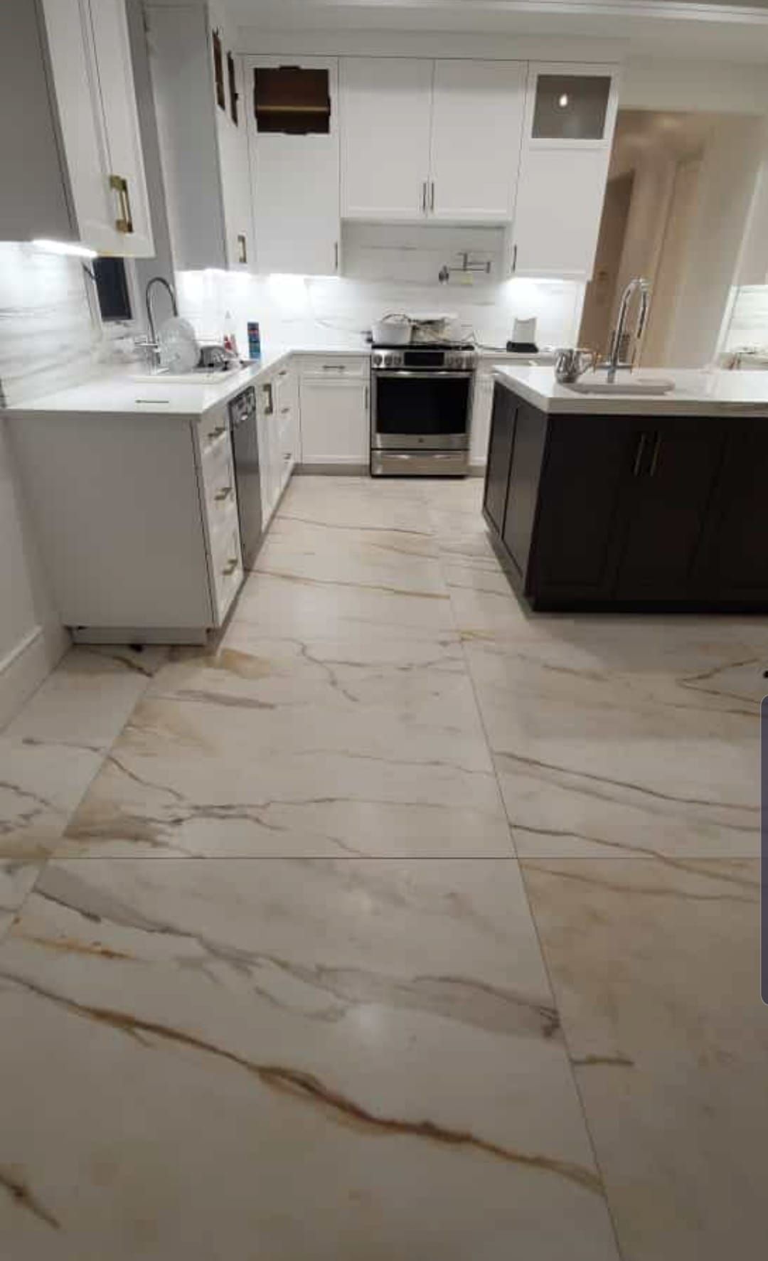 Modern white kitchen with marble-look tile floor, dark island cabinets, and stainless steel stove.