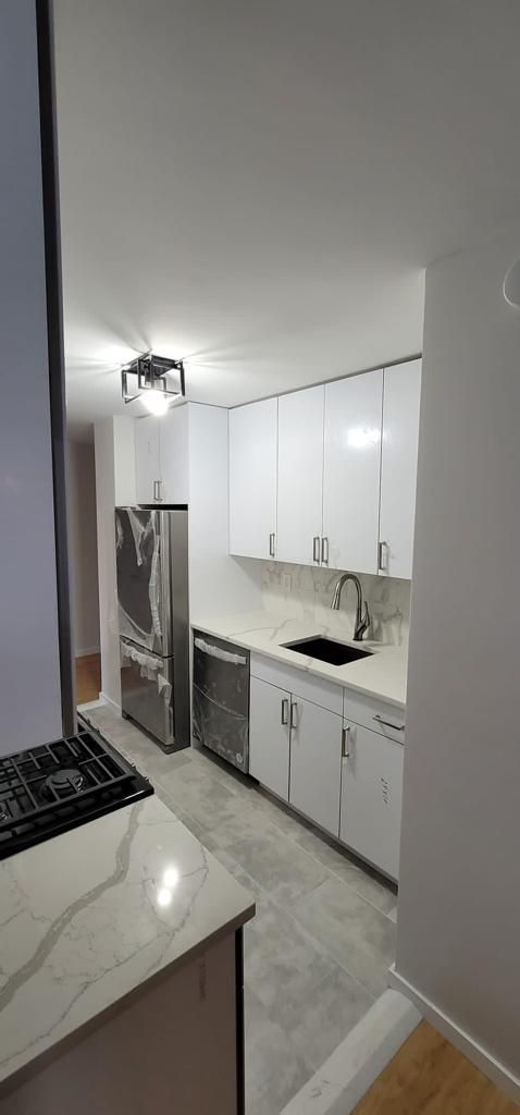 Narrow modern kitchen with white cabinets, black countertops, and a lit ceiling fixture.