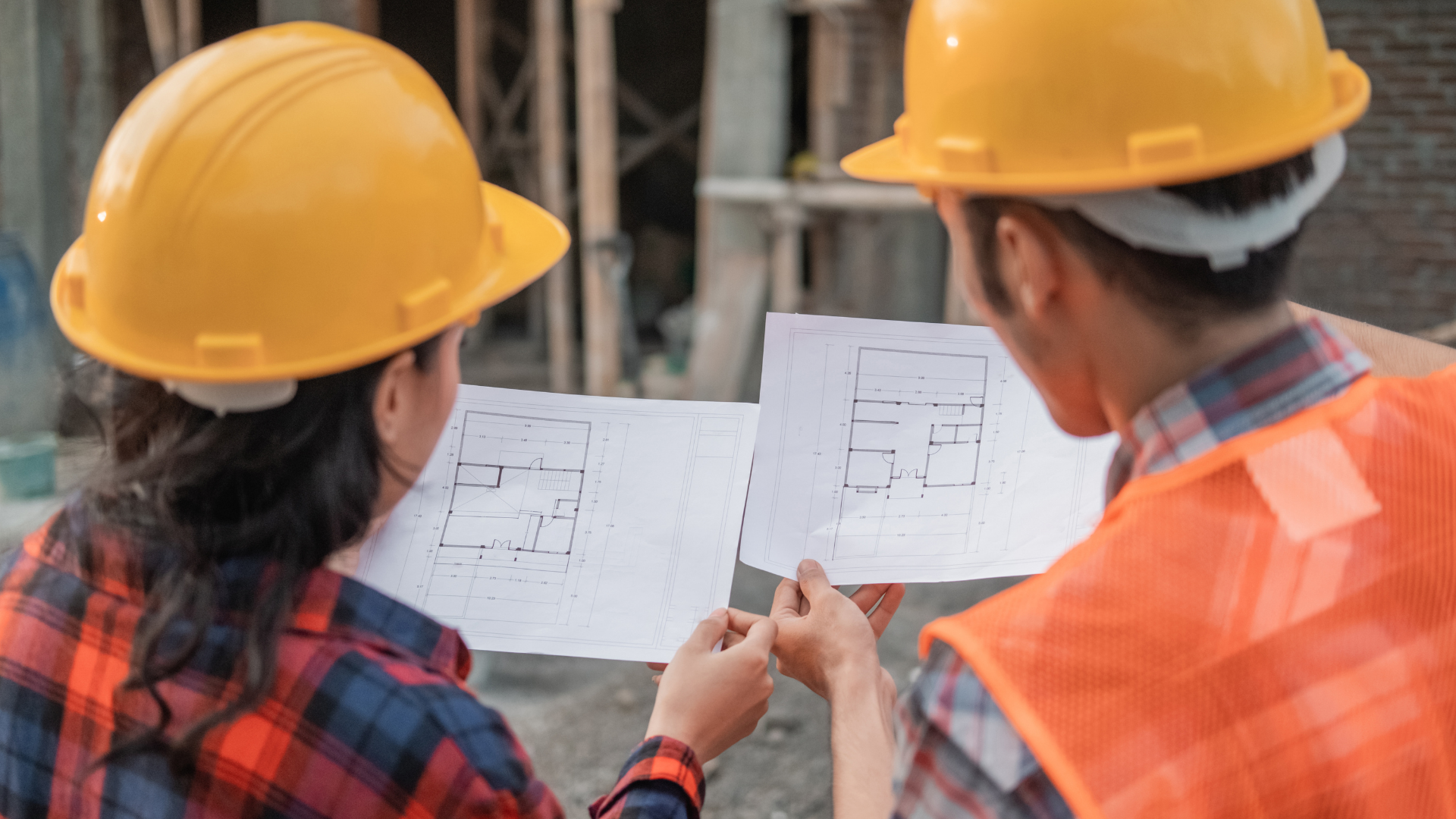 Two construction workers in yellow hard hats reviewing blueprints at a building site