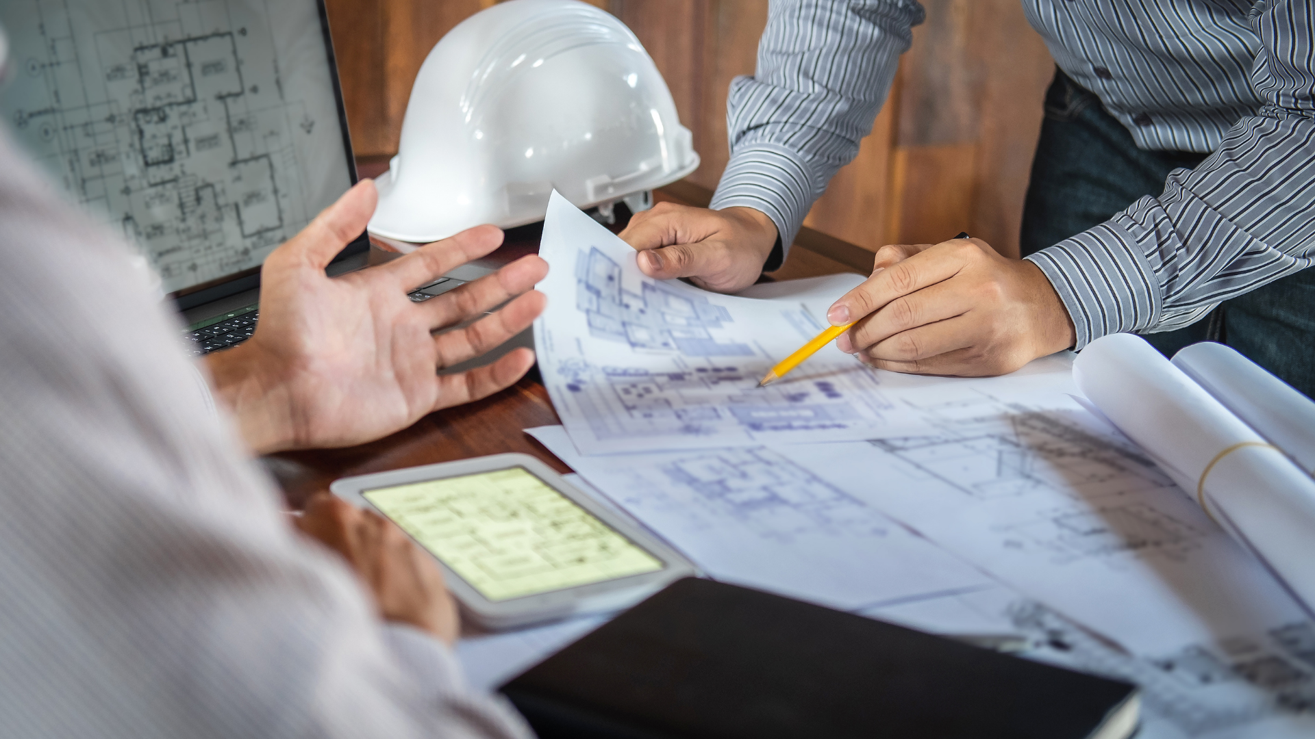 People reviewing blueprints and notes at a construction meeting, with a hard hat on the table
