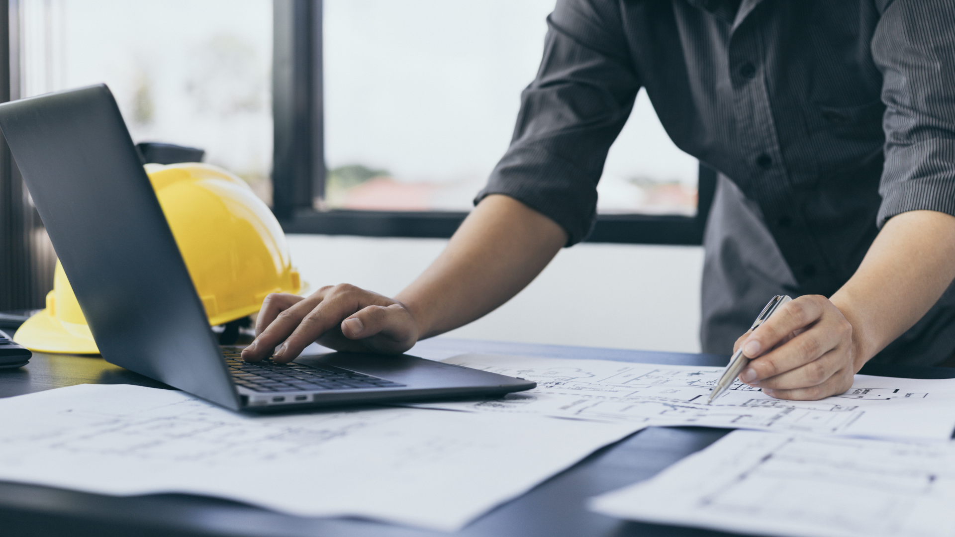 Person working at a desk with laptop, hard hat, and architectural plans