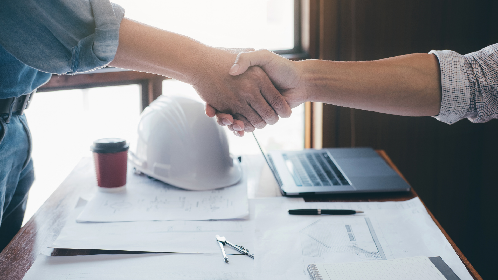 Two people shaking hands across a desk with a laptop, hard hat, and papers in view