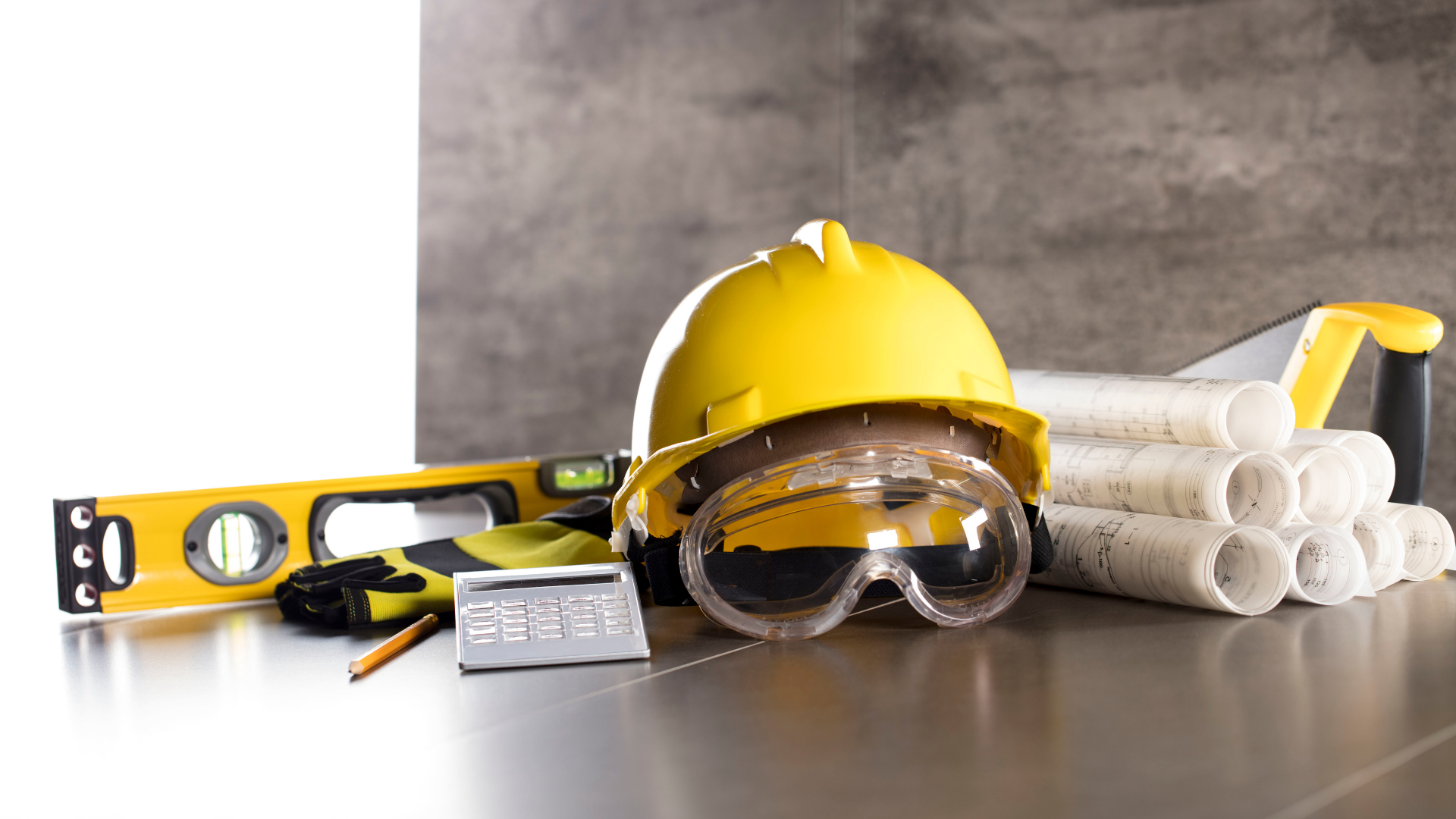 Yellow hard hat, safety goggles, level, and rolled blueprints on a table