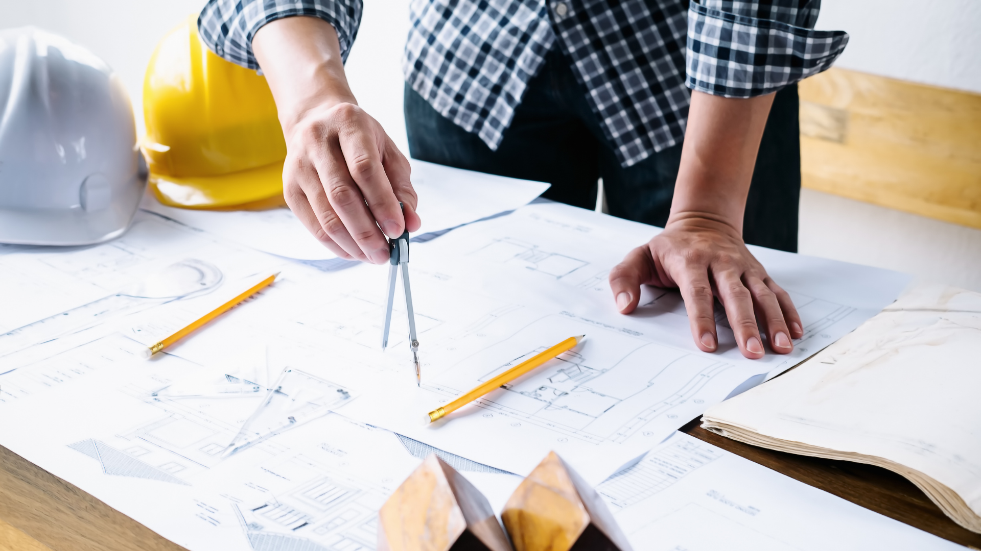 Hands marking blueprints on a desk with pencils and a yellow hard hat nearby