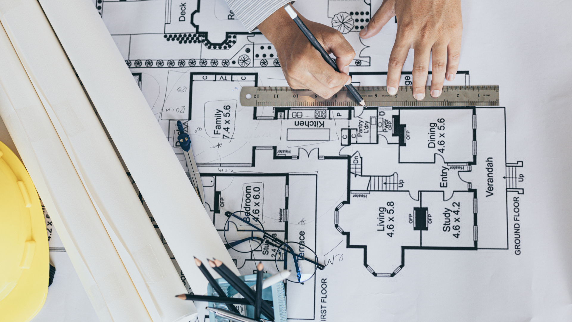 Hands marking measurements on a floor plan with a ruler and pen beside drafting tools.