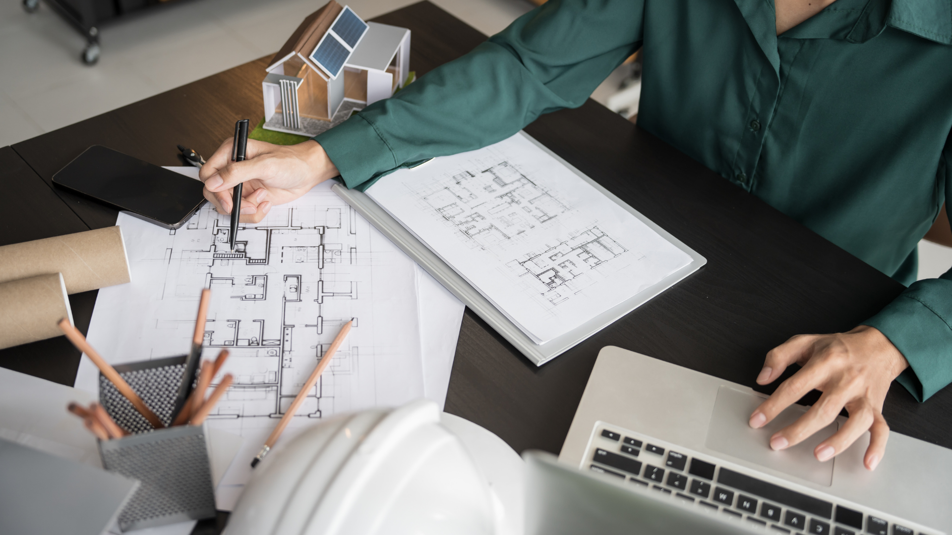 Person working on house plans at a desk with laptop, ruler, pencil, and architectural models