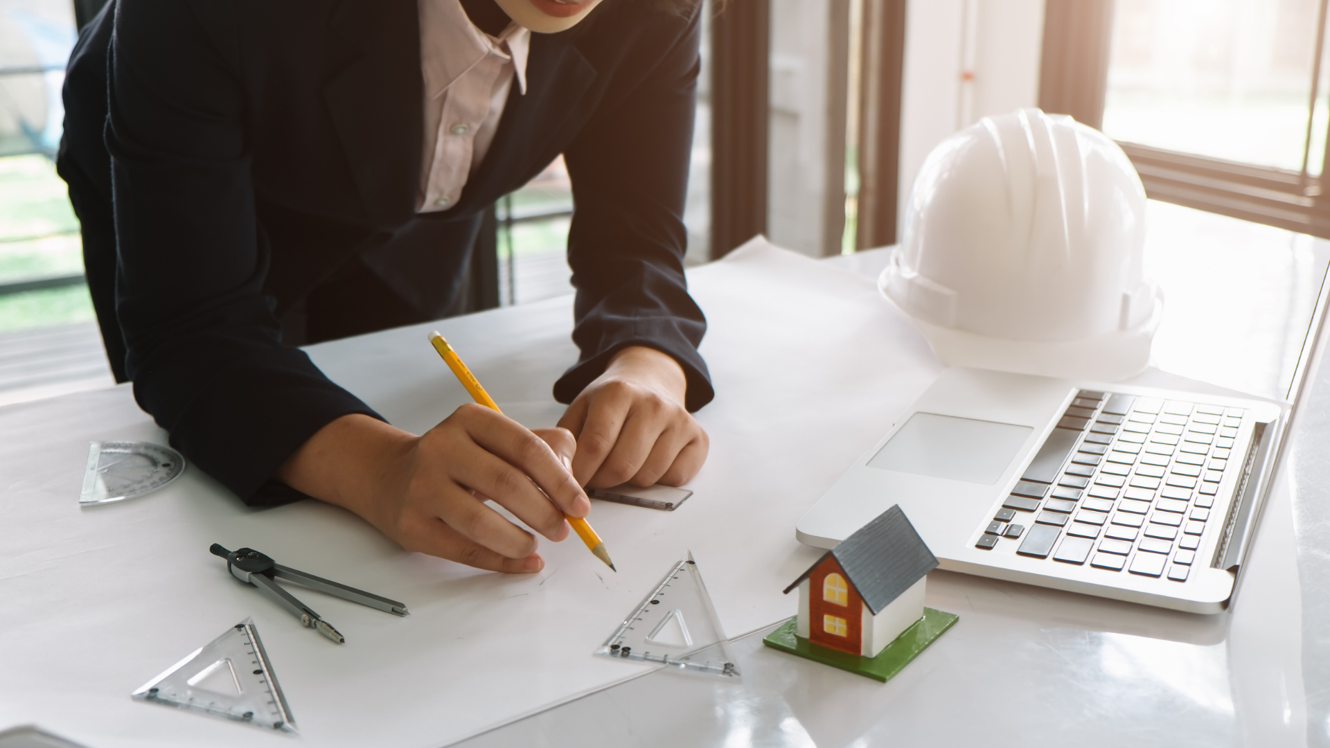 Engineer drafting house plans with a pencil on a desk, with ruler, model house, hard hat, and laptop.