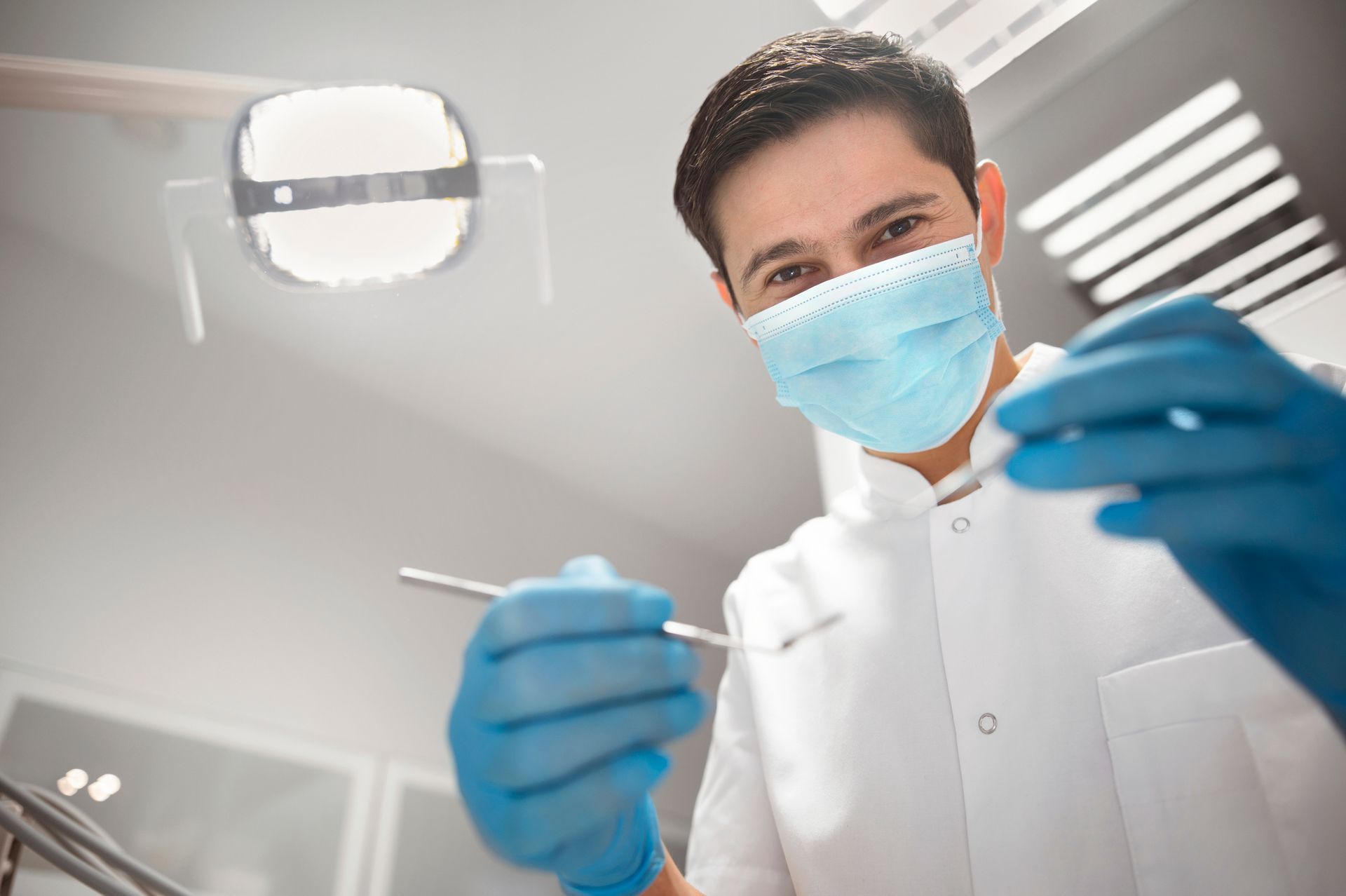 A dentist in gloves and mask holding tools under a bright light in a clean, modern dental clinic
