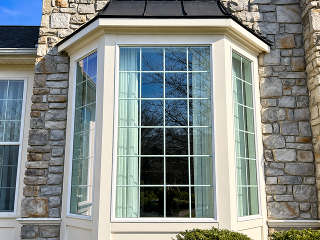 Bay window with white trim on a stone house, reflecting trees in the glass.