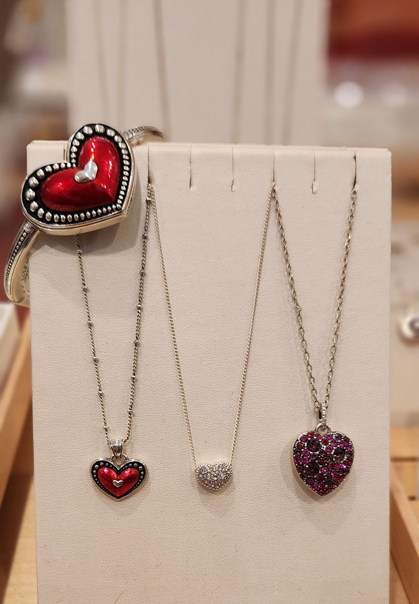 Three heart-shaped pendant necklaces displayed on a beige jewelry stand, one with a red heart ring beside it