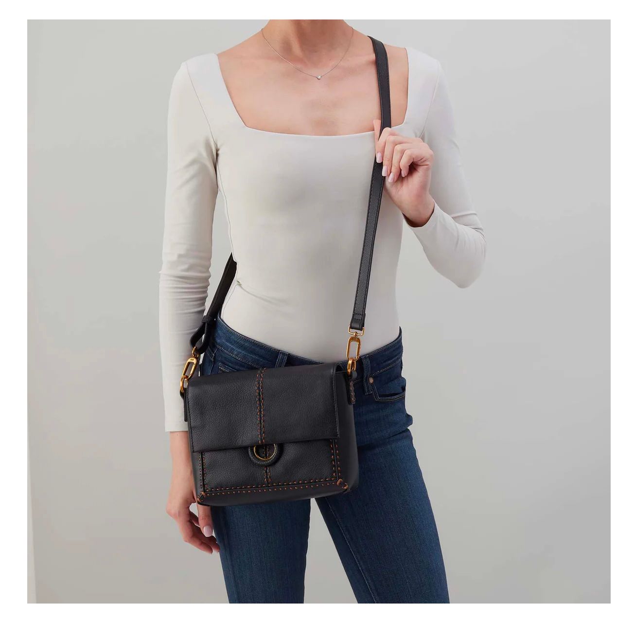 Woman wearing a beige top and jeans with a black crossbody bag against a gray background
