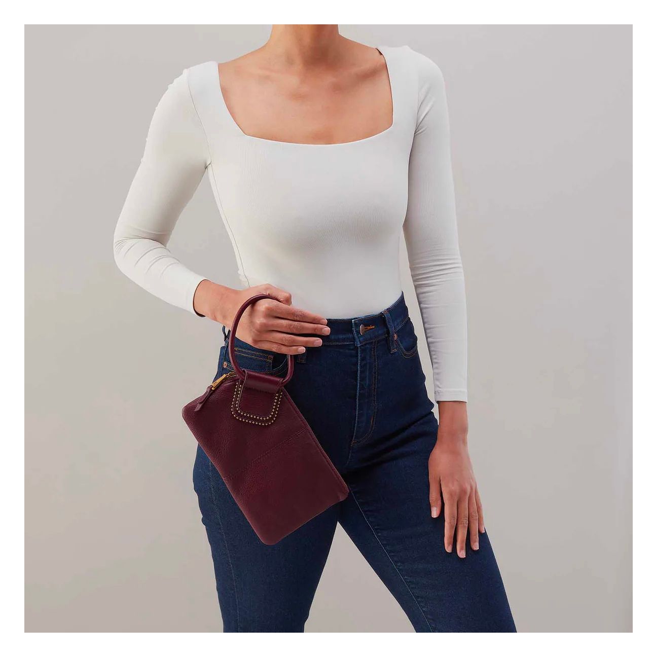 Person in a white long-sleeve top and dark jeans holding a burgundy handbag against a gray background
