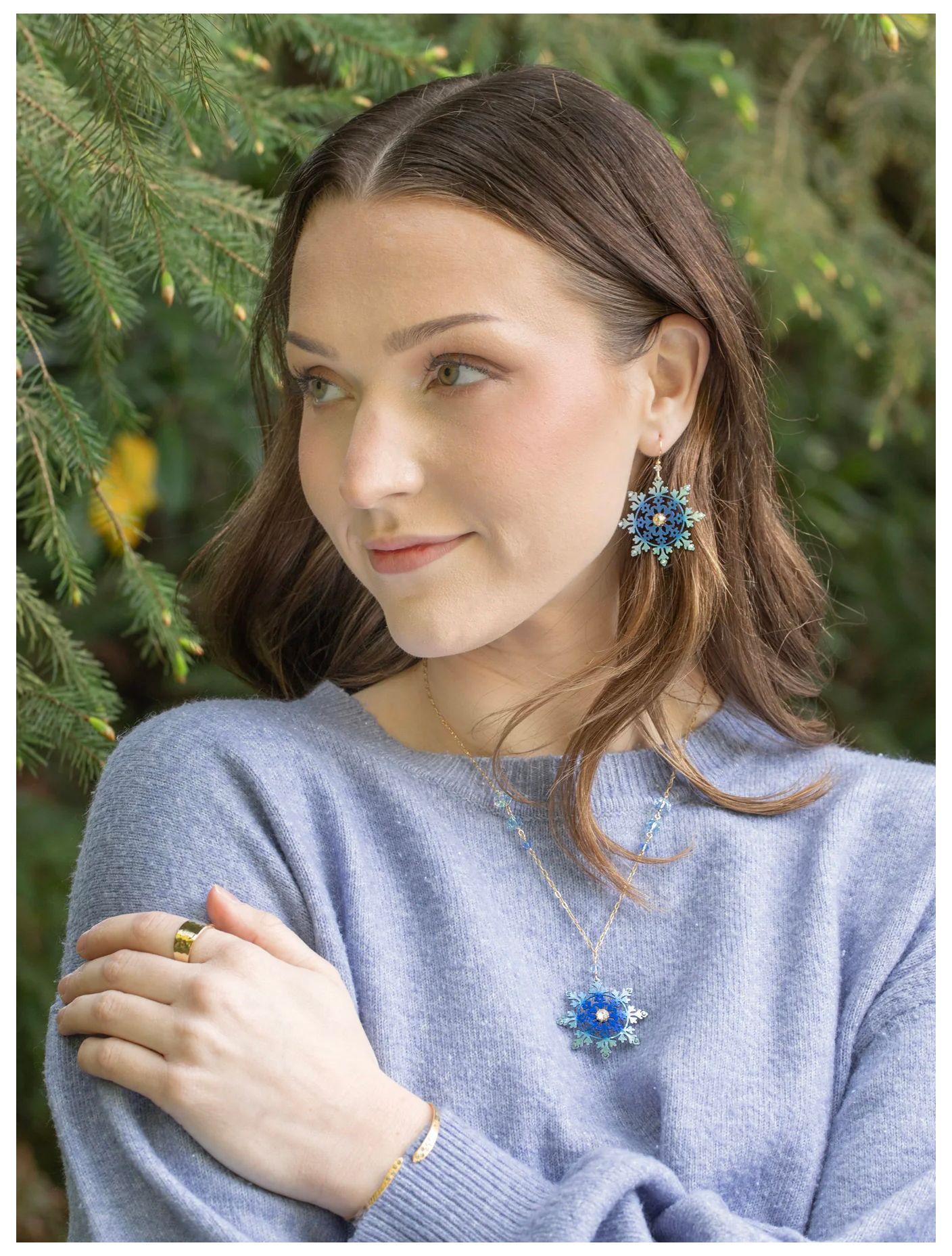 Woman in a light blue sweater wearing blue star earrings and necklace, standing outdoors by green foliage.