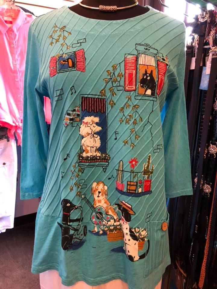 Teal long-sleeve shirt with colorful cat and window illustrations hanging in a clothing store