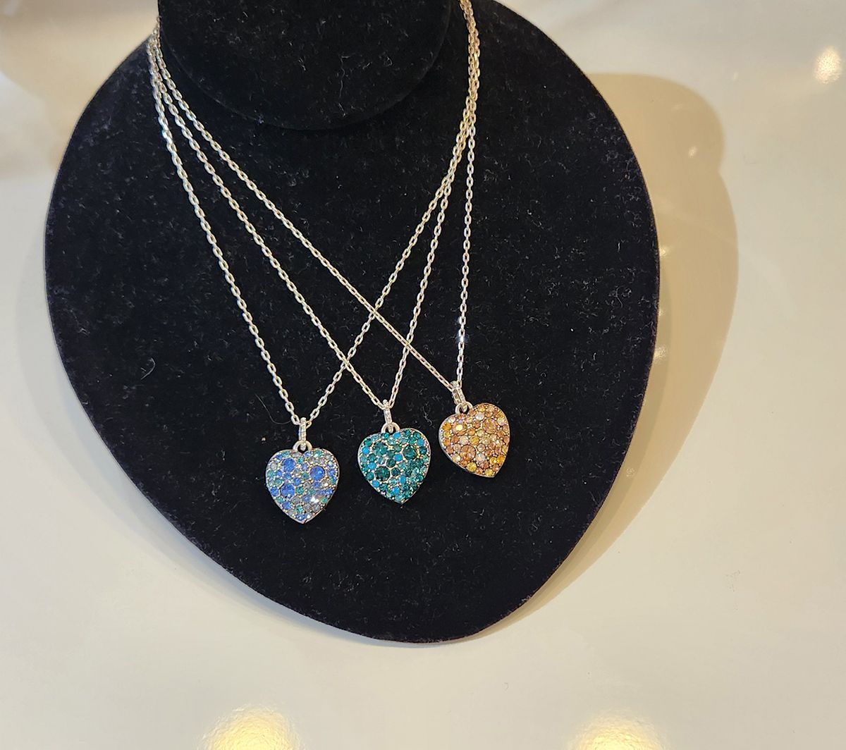 Three heart-shaped gemstone necklaces on a black display bust, in blue, teal, and amber.