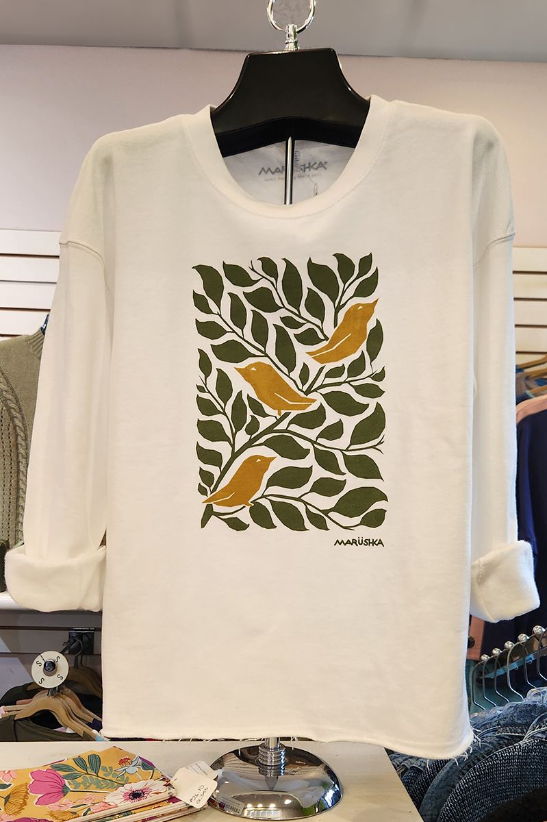 White long-sleeve shirt with green leaf and orange bird graphic on a hanger in a clothing store.