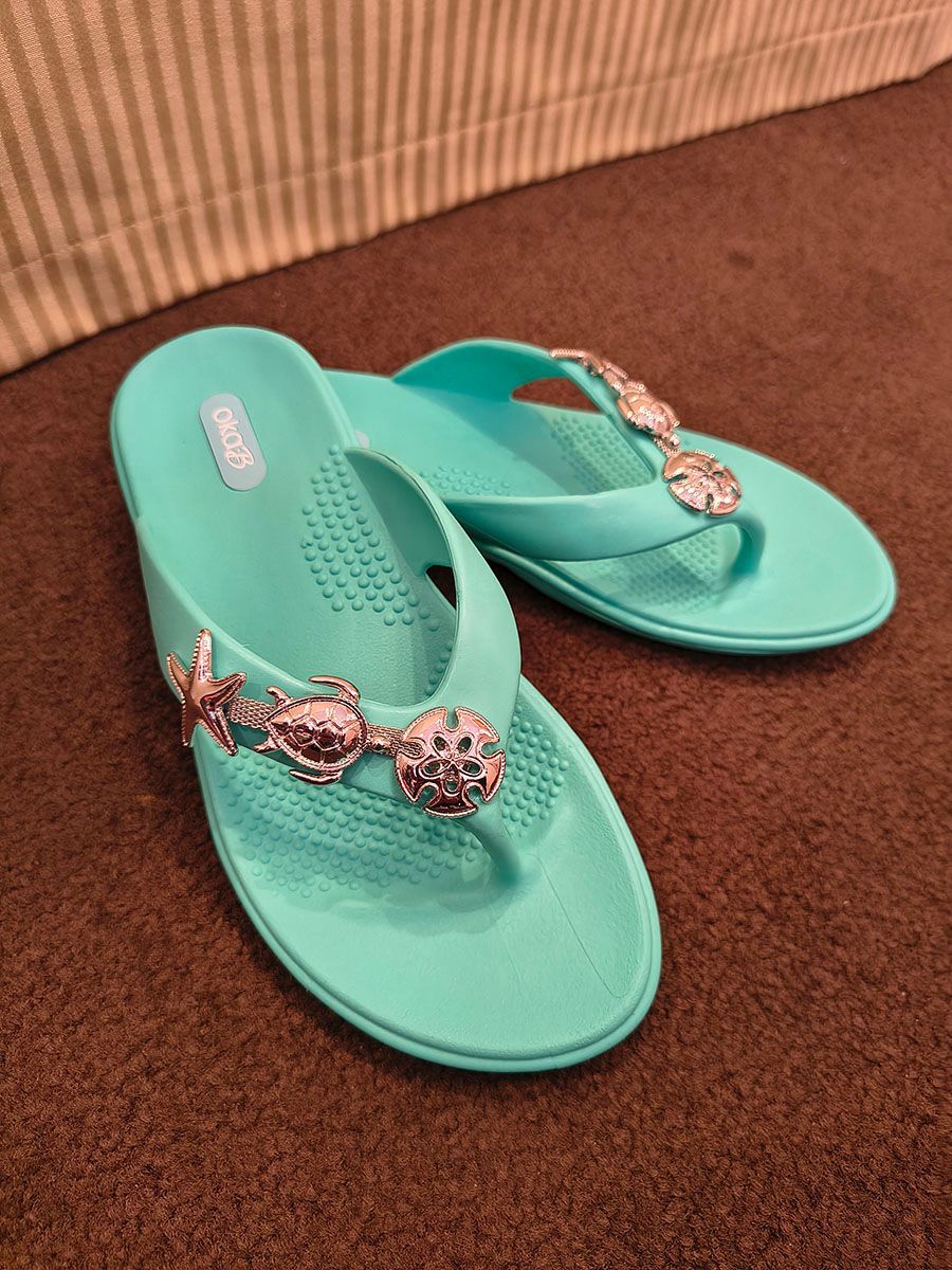 Turquoise flip-flops with jeweled straps on a brown carpet near a striped cushion