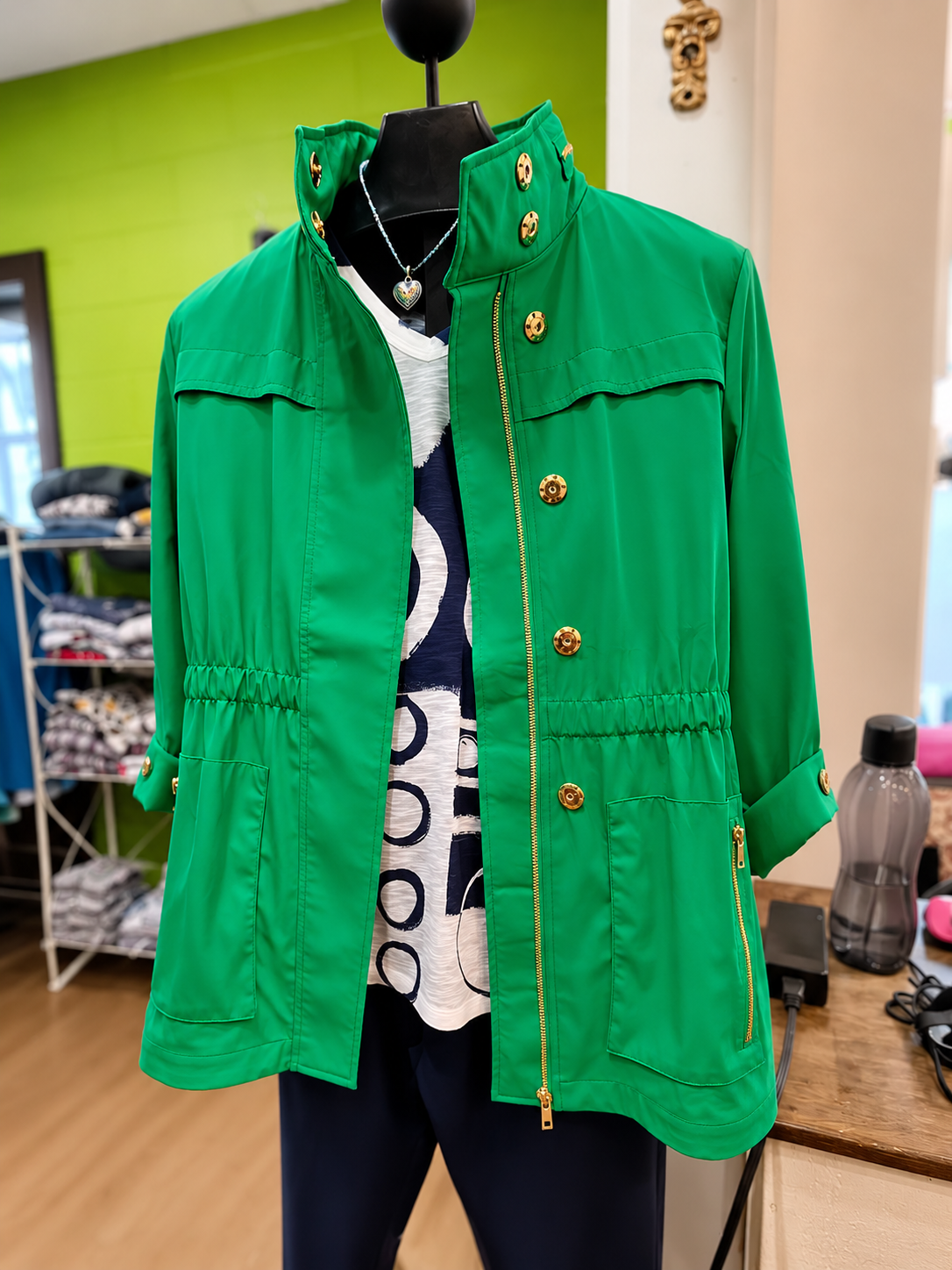 Green button-up jacket over a white patterned top on a black mannequin in a clothing store.