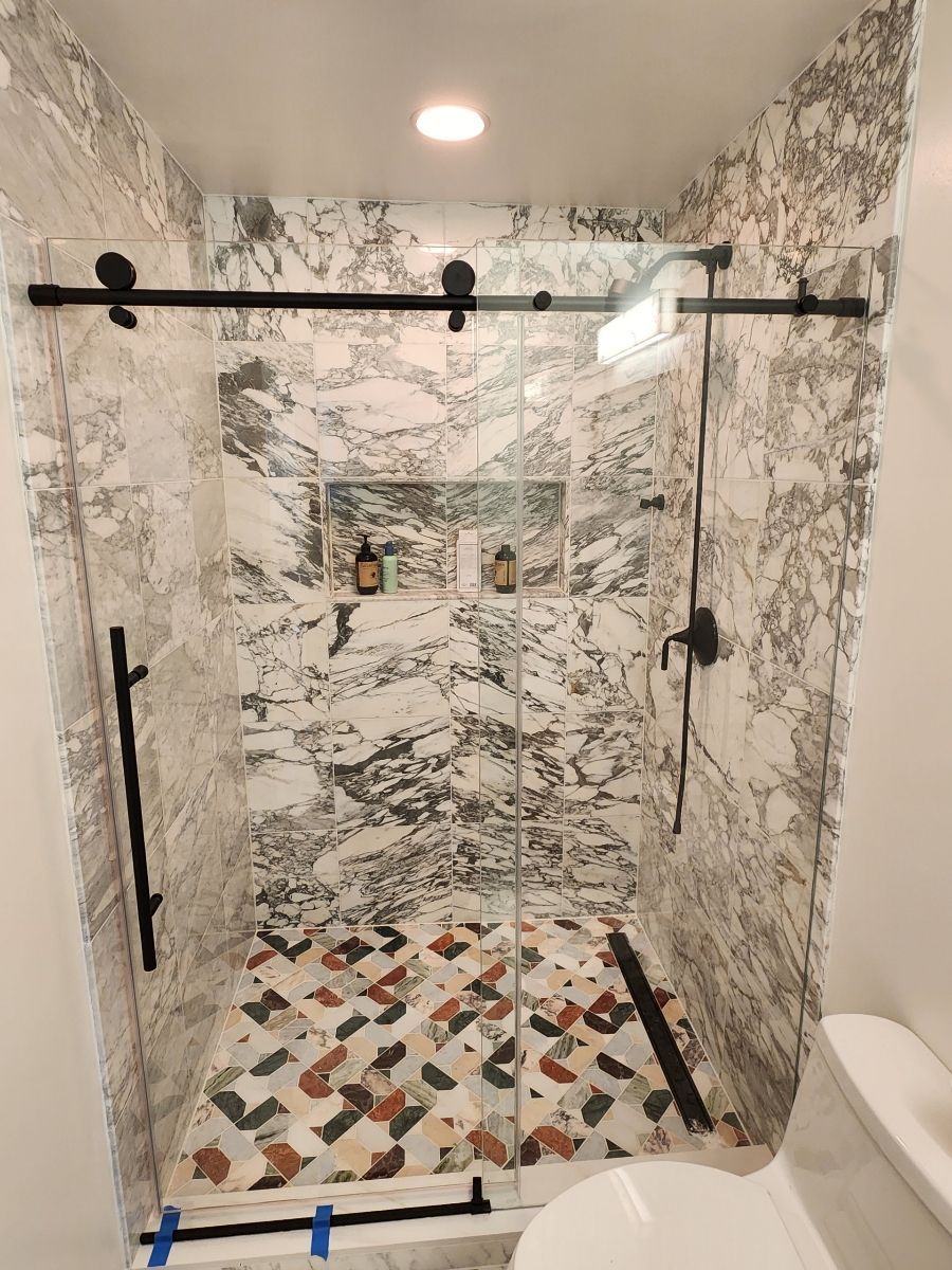 Shower with marble walls, patterned floor tiles, black hardware, and glass sliding door.