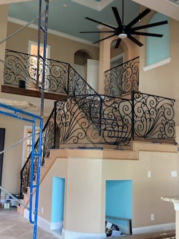A grand staircase features ornate black iron railings, a blue ceiling, and a large ceiling fan under construction.