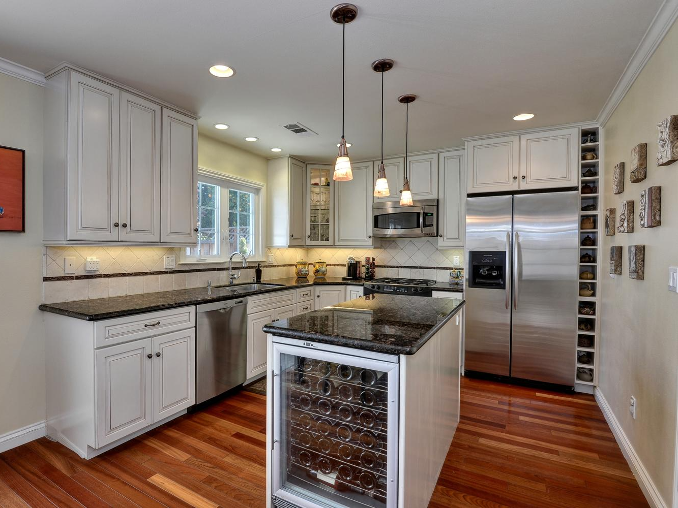 Kitchen with white cabinets, dark countertops, island with wine fridge, stainless steel appliances, and wood floors. Andrea Lim | Practical Vanity Interiors | Granite Bay, CA | Interior Design