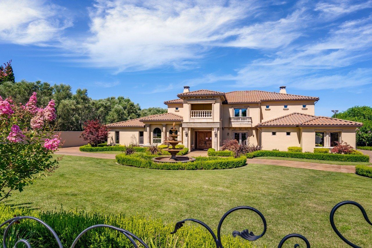 Luxurious beige mansion with tile roof, fountain, and manicured lawn under a blue sky.