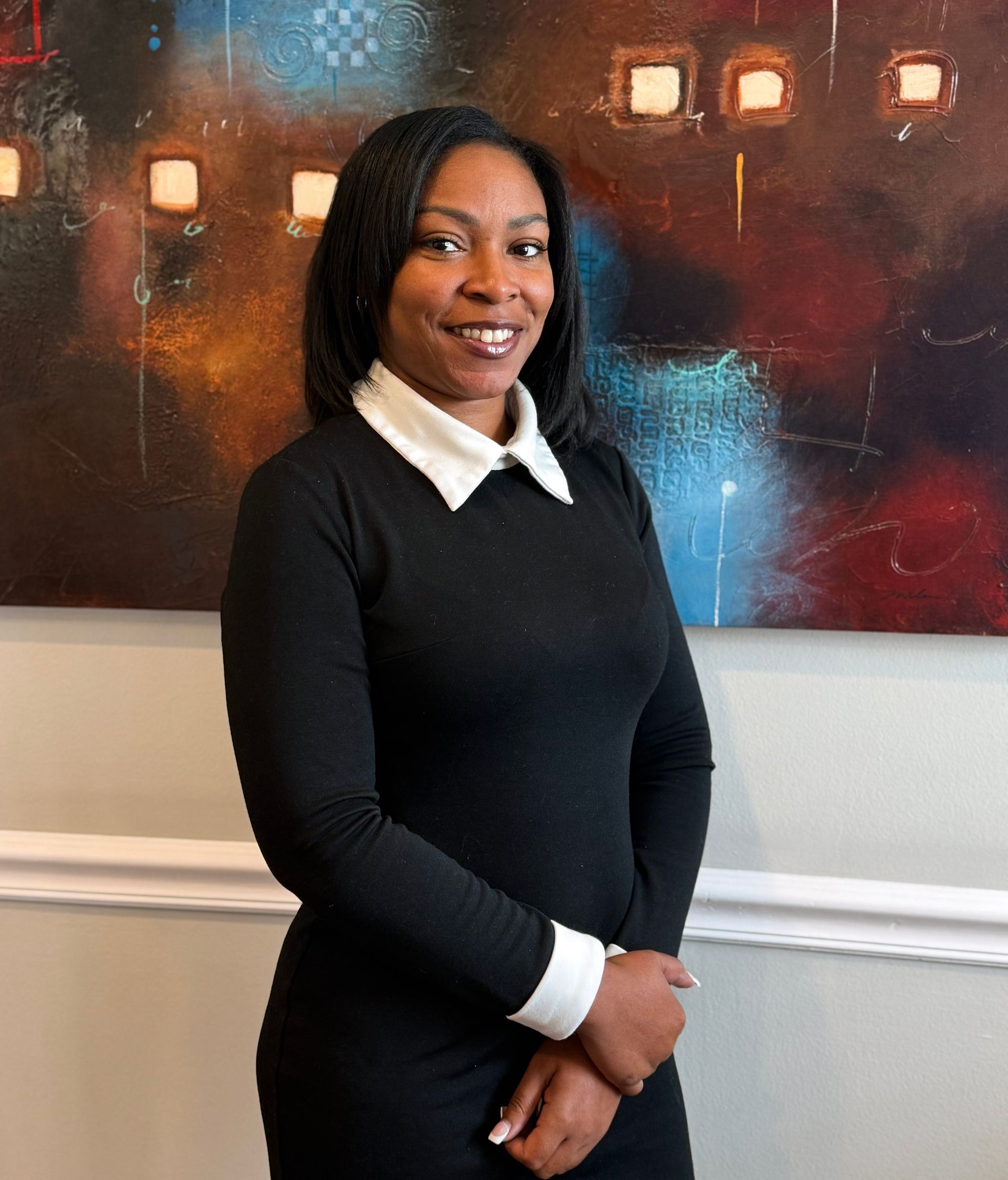 Woman in black dress with white collar and cuffs, smiling, posing in front of abstract art.