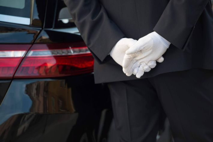 Tre uomini in abito nero sono in piedi accanto a un carro funebre argentato di fronte a un edificio con gradini.