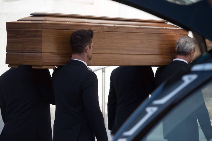 Tre uomini in abito nero sono in piedi accanto a un carro funebre argentato di fronte a un edificio con gradini.