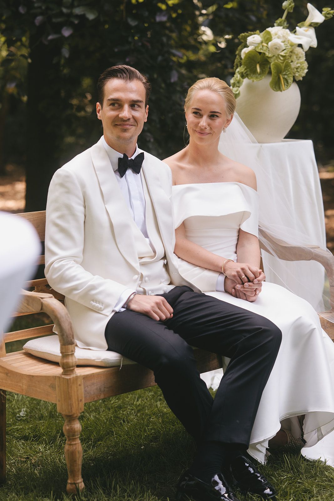 Bruidspaar op de bank. Bruidegom in een witte smokingjas, zwarte vlinderstrik en zwarte pantalon. De bruid in een witte trouwjurk met strapless schouders