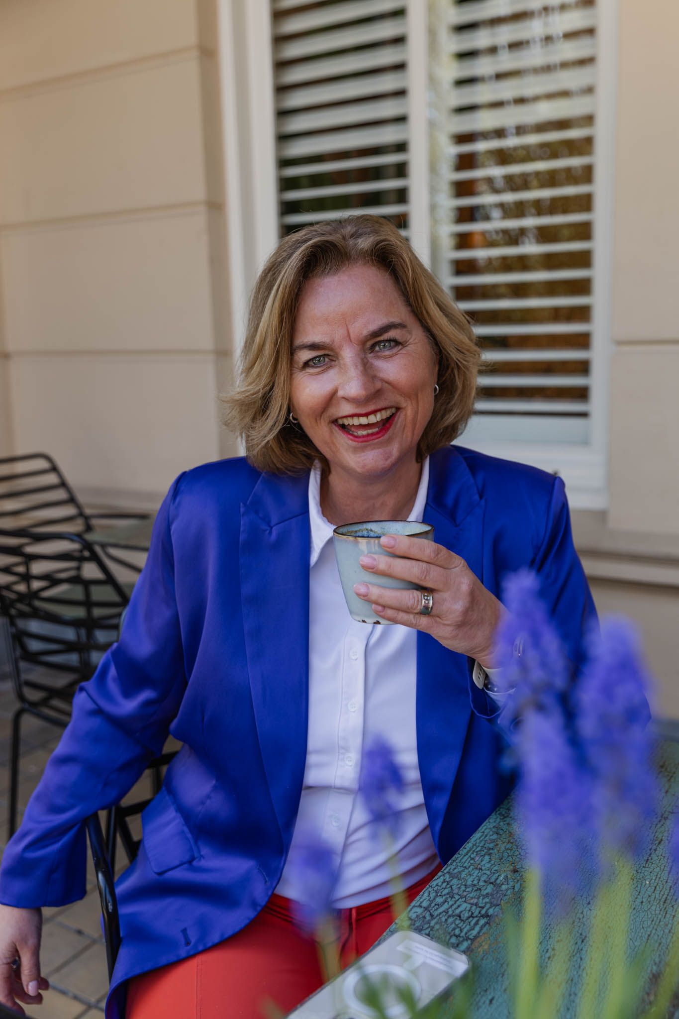 Foto gemaakt door https://marlotbakker.nl/ Trouwambtenaar in prive houding afgebeeld. Kopje koffie in de hand, blauwe blazer, witte blouse en oranje broek aan. Zittend aan een tafeltje.