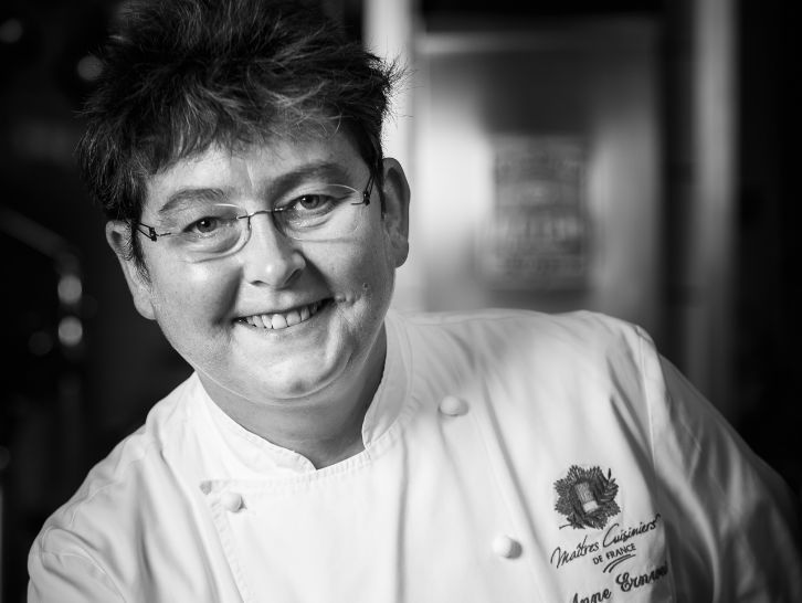 Une femme portant des lunettes et une veste de chef sourit sur une photo en noir et blanc.