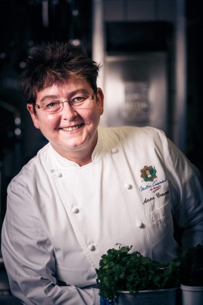 Une femme en uniforme de chef sourit et tient une plante en pot.
