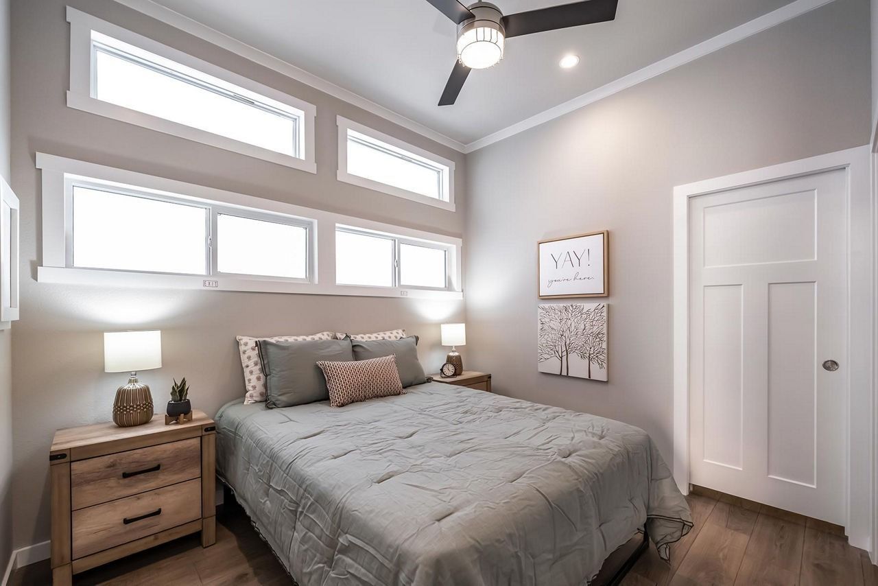 Bright modern bedroom with a gray bed, wood nightstands, high windows, and a ceiling fan