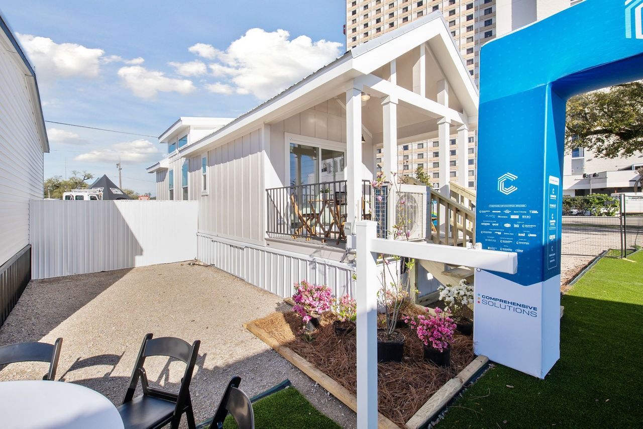 White tiny house display with porch, blue sign, and flower bed at an outdoor event